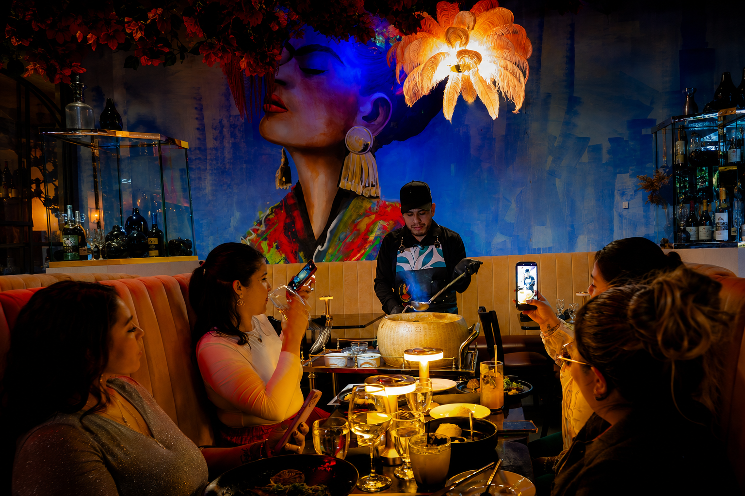 Guests enjoy an interactive fine-dining experience as a chef prepares a signature dish tableside in a stylish restaurant. Warm golden lighting, vibrant wall art, and elegant décor create a luxurious atmosphere while diners capture the moment on their smartphones. The colorful mural and dramatic chandelier add a modern, upscale ambiance to the scene.